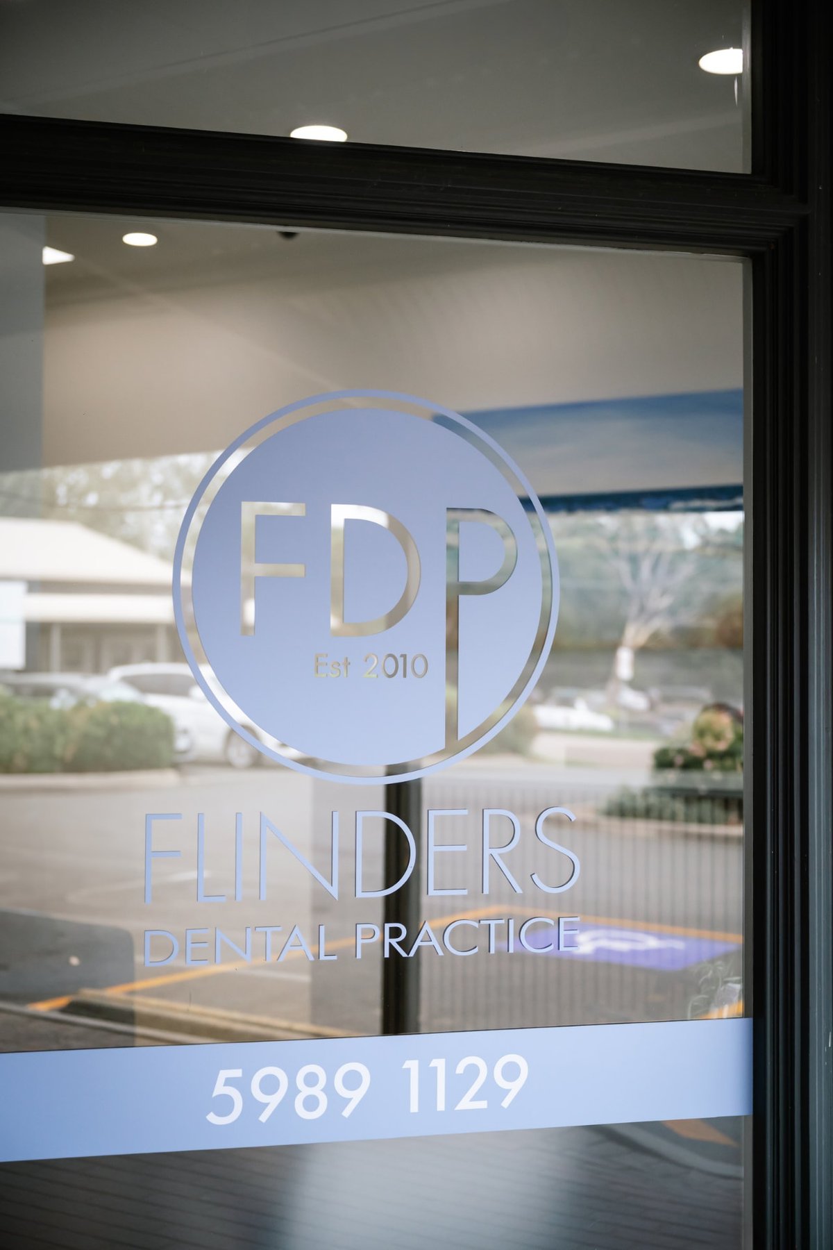 The glass front door of Flinders Dental Practice, with the practice phone number on the glass and the car park behind